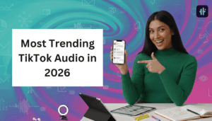 Smiling woman in a green sweater sits at a desk, pointing at a smartphone displaying a social media app. A large text overlay reads “Most Trending TikTok Audio in 2026” against a colorful abstract background with digital icons. A tablet, notebook, and small ring light are on the desk.